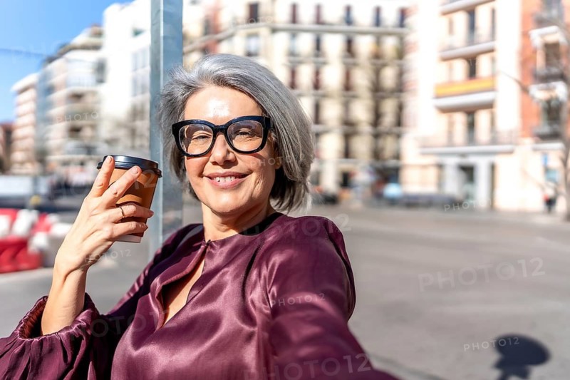 Happy mature woman with gray hair and glasses smiling at camera, holding takeaway coffee cup while enjoying a sunny stroll through a vibrant european city street