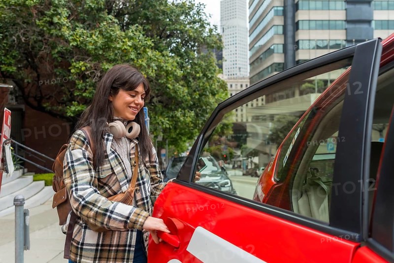 Young woman opens the door of a red rideshare car in san francisco, stepping onto a busy downtown sidewalk, commuting with backpack and headphones, smiling and on the go