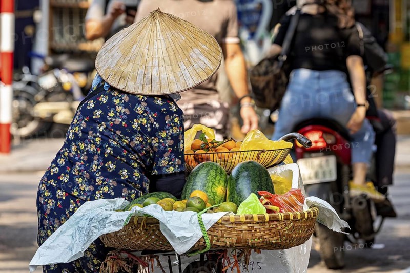 Vendor in traditional dress offering various fruits including watermelon on the street, woman selling fruits in the streets of Hanoi Vietnam
