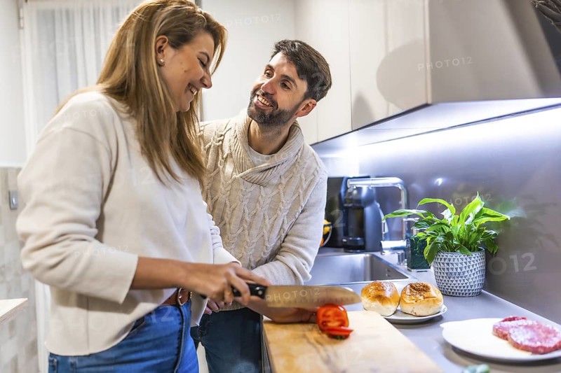 Smiling couple preparing dinner together, slicing tomatoes for burgers in their contemporary kitchen