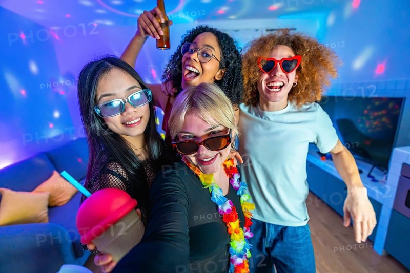 Cheerful young friends are enjoying a home disco party, taking a selfie with colorful lights and drinks, wearing funny sunglasses and having fun
