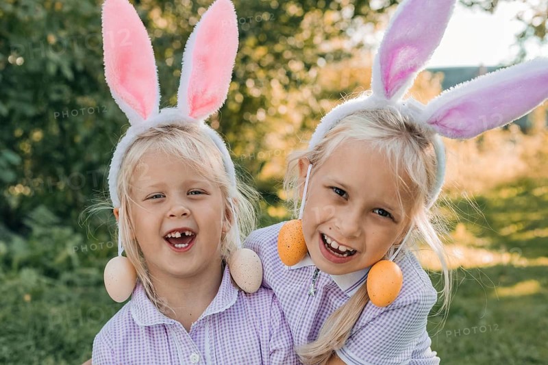 Two joyful children with bunny ear headbands celebrate Easter outdoors, laughing and playing with decorative eggs. A heartwarming scene of childhood and festivity
