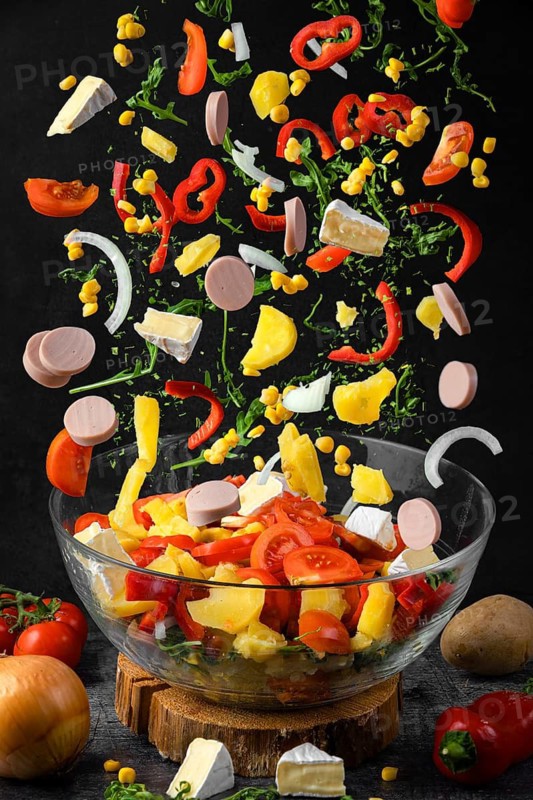 Salad ingredients, such as peppers, tomatoes, sausage, corn, arugula and potatoes fall a glass bowl, black background