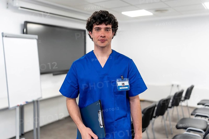 Young man wearing blue medical scrubs and an id badge, holding a clipboard while standing in a training classroom, representing healthcare education and professional development