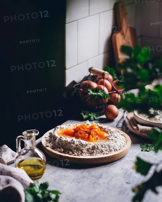 Rustic kitchen scene showcasing a mound of flour with eggs and vibrant yolks, alongside a glass jar of olive oil and scattered fresh herbs. Perfect for baking and cooking themes