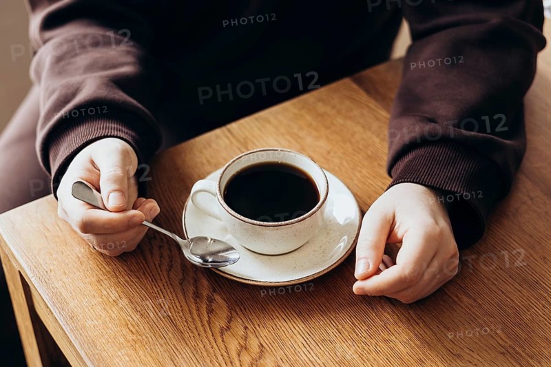 Close up of hands holding a spoon near a cup of coffee on a wooden table in a cafe. The scene conveys a cozy and relaxed atmosphere, perfect for coffee lovers and peaceful mornings