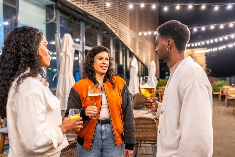 Three multi-ethnic friends drinking beer and talking standing on a bar terrace at night