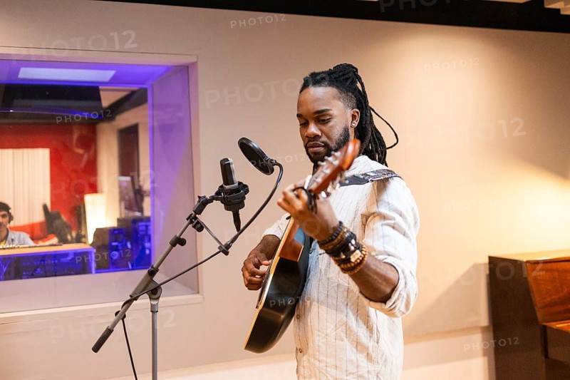 A musician with headphones plays an acoustic guitar in a professional recording studio, using state-of-the-art microphones with a sound technician visible in the control room