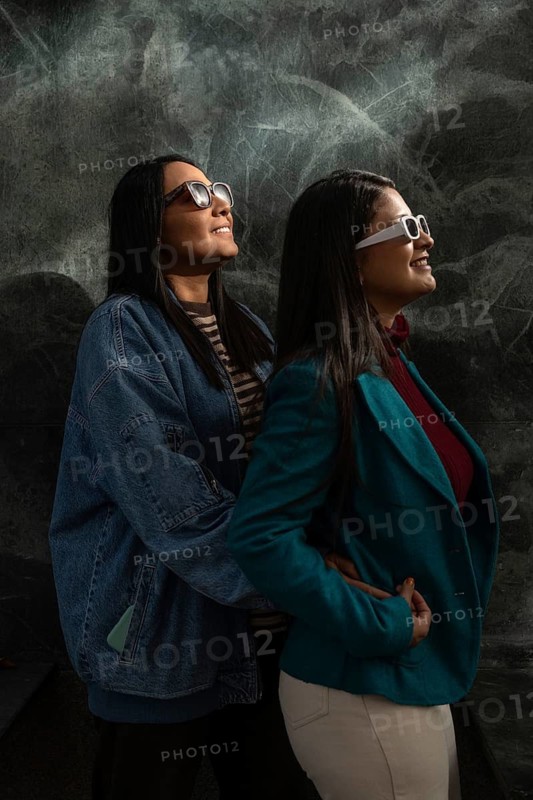 A cheerful Latin lesbian couple wears stylish sunglasses while embracing. Both women smile warmly, sharing a joyful moment together against a textured background