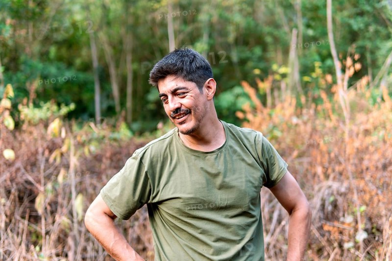 A middle-aged Colombian man takes a joyful break amid farm work in the rural Colombian countryside, surrounded by lush greenery and under the soft daylight. His content expression reflects a moment of relaxation from physical labor