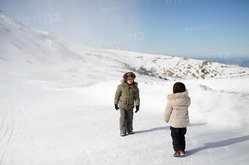 Two children wearing winter jackets play in the snow at Sierra Nevada Ski Resort. The clear sky and snow-covered landscape create a perfect winter scene