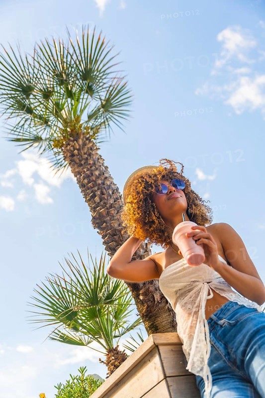 Vertical low angle view of a sensual brunette woman with curly hair wearing sunglasses and hat, drinking juice under palm tree