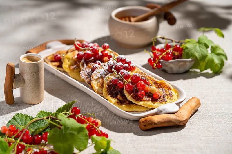 From above, a delicious stack of pancakes topped with vibrant red currants sits on a white platter. The background features a linen cloth, enhancing the rustic appeal