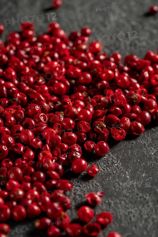 A close-up view of vibrant red peppercorns heaped on a textured dark slate surface, highlighting the shiny and textured exterior of each peppercorn