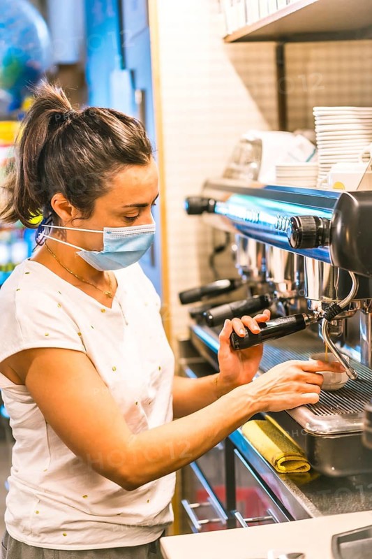 Photo session with a waitress with a face mask in a bar. New normality, security measures after the coronavirus pandemic, social distance, covid-19. Preparing a cut of coffee with milk with the machin