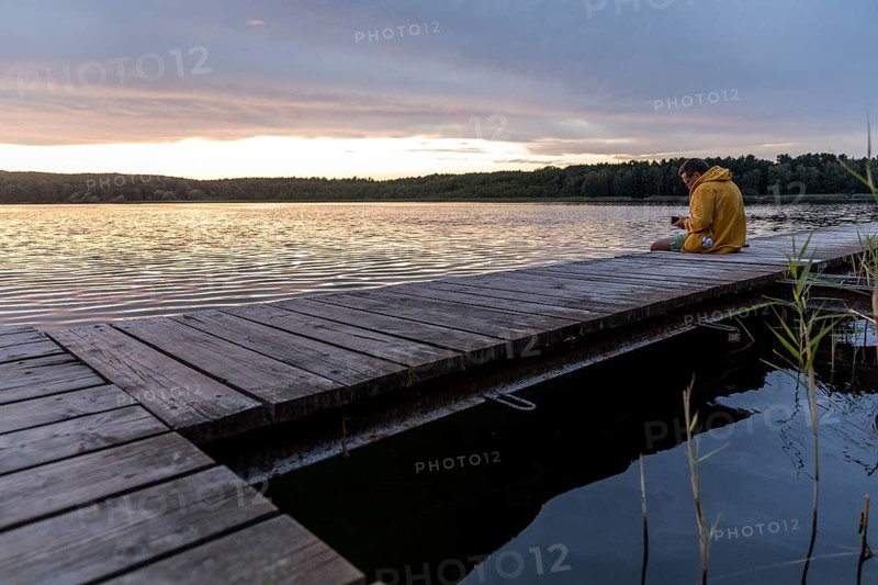 Man sitting on the bathing jetty and looking at his mobile phone, evening mood at the Großer Glubigsee near Wendisch Rietz, Oder-Spree district, Brandenburg, Germany