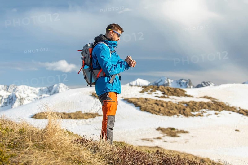 A man dressed in vibrant outdoor gear stands on a snowy landscape in the Pyrenees. With a backpack and sunglasses, he enjoys the breathtaking mountain views