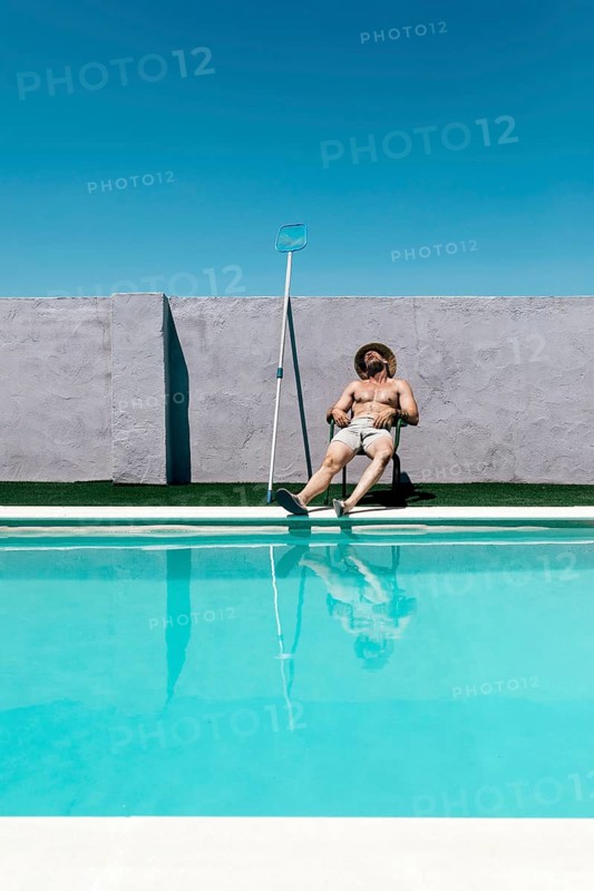 A shirtless man in shorts and a straw hat takes a nap, relaxing on a chair by a clear blue swimming pool, with pool cleaning equipment nearby, under a clear blue sky
