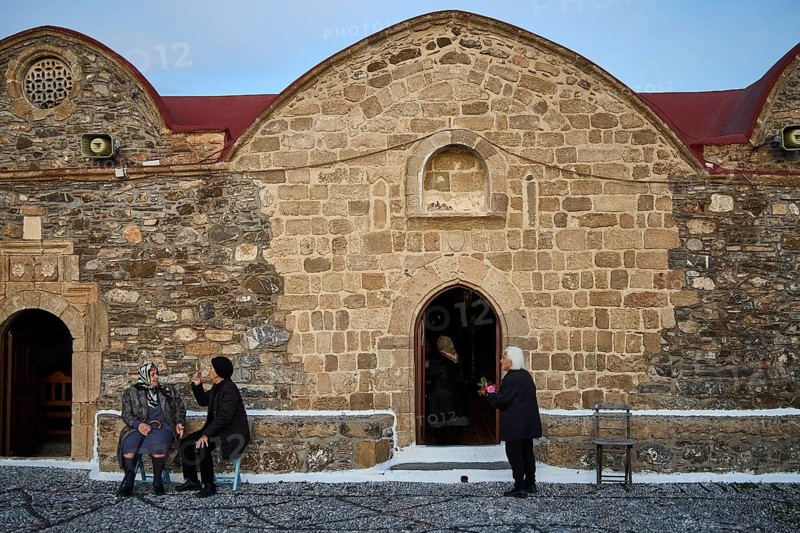 People in front of an old stone wall in conversation and on the pavement, Easter 2025, Church of the Dormition of the All-Holy God Bear, Kimisis tis Theotokou, Asklipio, Rhodes, Dodecanese, Greek Islands, Greece