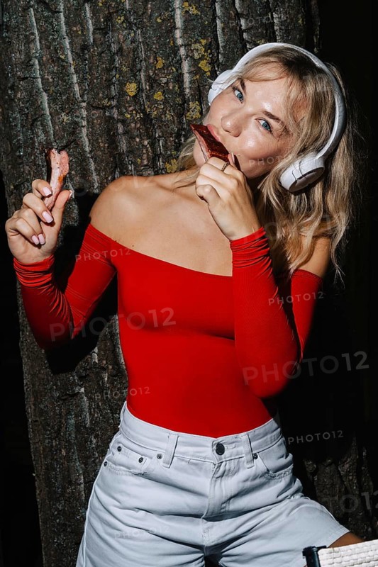 A woman in a vibrant red top and white shorts enjoys barbecue ribs while wearing headphones, set against a tree background. A blend of food, fashion, and leisure
