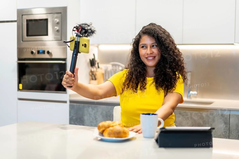 Happy latin female young vlogger recording online while eating breakfast sitting in the kitchen at home