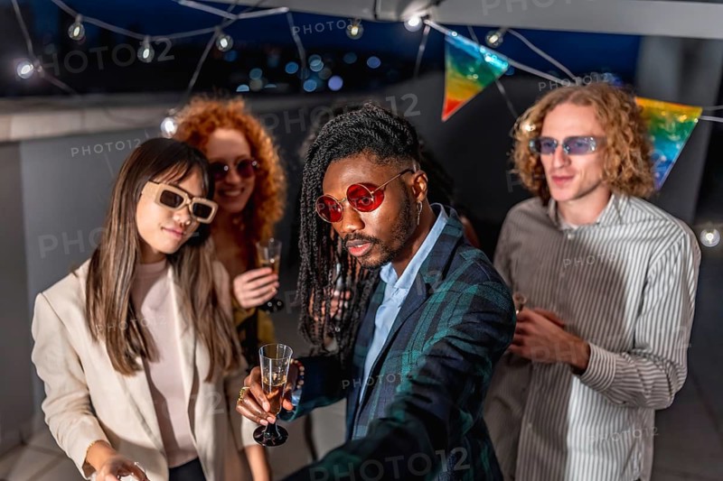 African stylish man taking selfie with multi-ethnic friends wearing elegant clothes and sunglasses during night party in a terrace at night