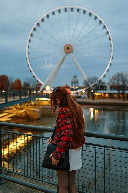 Back view of unrecognizable woman with red hair and a plaid jacket gazing at the illuminated Ferris wheel in Montreal. The cityscape hints at a chilly fall evening