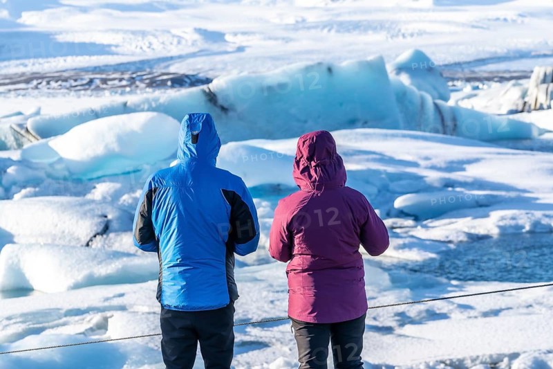 Two people are standing on a frozen lake, looking out at the ice. The man is wearing a blue jacket and the woman is wearing a purple one