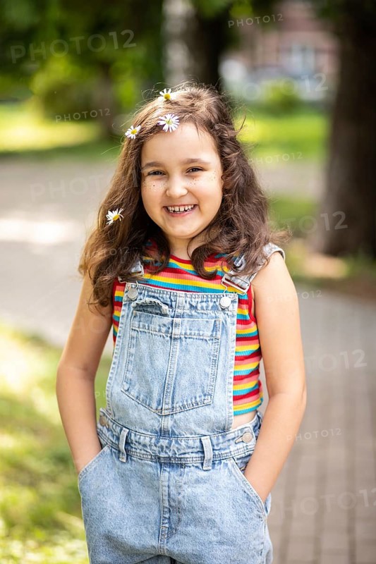 A cheerful little girl with long hair adorned with chamomile flowers smiles warmly at the camera. She stands outdoors, dressed in denim overalls, enjoying a sunny day