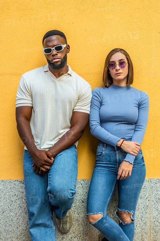 Vertical photo of two stylish multiracial people in casual denim clothes and sunglasses leaning on an urban wall