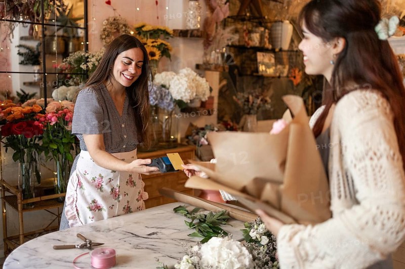 Two women engage in a friendly transaction at a vibrant florist shop. Surrounded by colorful flowers, they exchange a bouquet for payment, showcasing small business charm