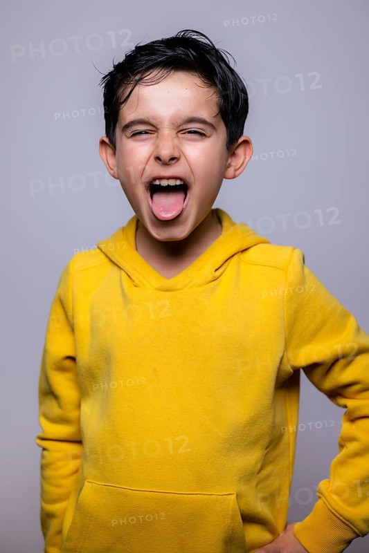 Young boy in a vibrant yellow hoodie playfully sticking out his tongue while expressing excitement or joy. The neutral background emphasizes his animated expression and attire