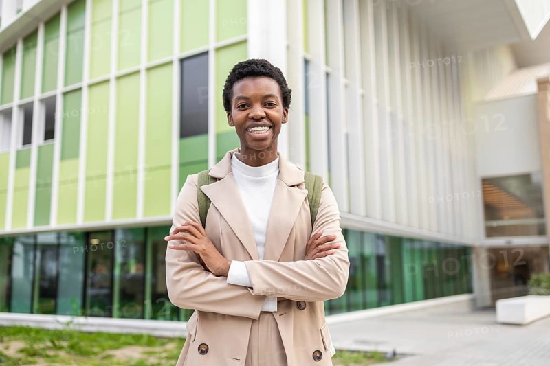 Confident black woman with short hair stands in front of a modern urban building, exuding empowerment and style She wears a beige outfit and carries a backpack