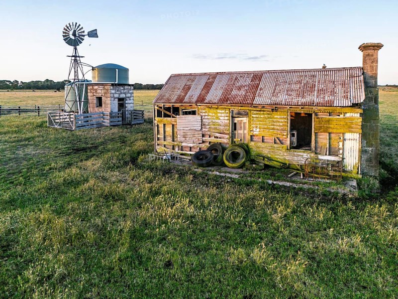 Weathered wooden farmhouse with a rusted roof, surrounded by grassy fields in Victoria, Australia. Adjacent stands a traditional metal windmill with a storage tank, capturing countryside charm