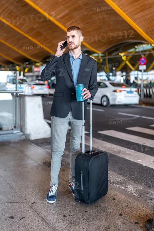 Businessman talking on the phone and holding a coffee while waiting at the airport with his luggage
