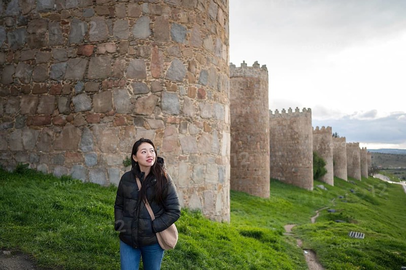 An Asian female tourist strolls along Avila's ancient medieval walls. The scene captures cultural exploration and architectural beauty
