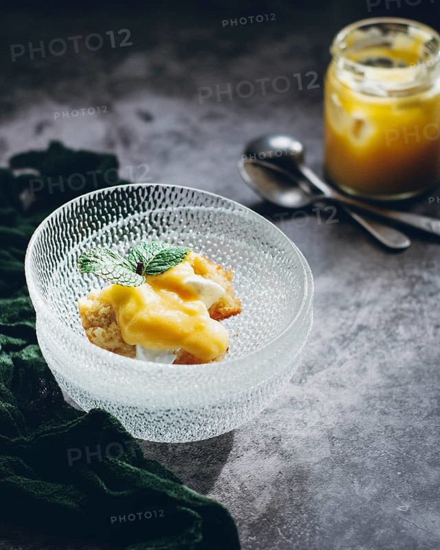 A delectable lemon curd cake, topped with a fresh mint leaf, served in an elegant glass dish. Perfectly styled, this dessert offers a delightful burst of citrus flavor