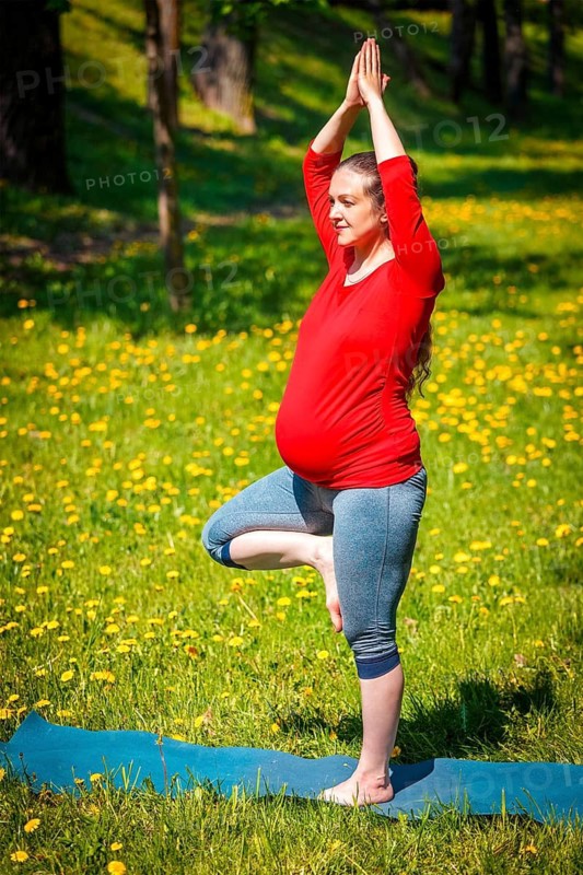 Pregnancy yoga exercise, pregnant woman doing asana vrikshasana tree pose outdoors on grass lawn with dandelions in summer