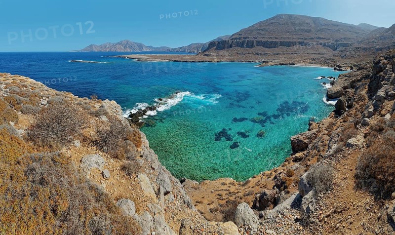 Panoramic views of a rocky coastline with clear turquoise waters and distant mountains under a clear sky, hike to the ancient Dorian city of Vrougounda and the cave church of St John Vrykountas, Avlona, Karpathos, Greece