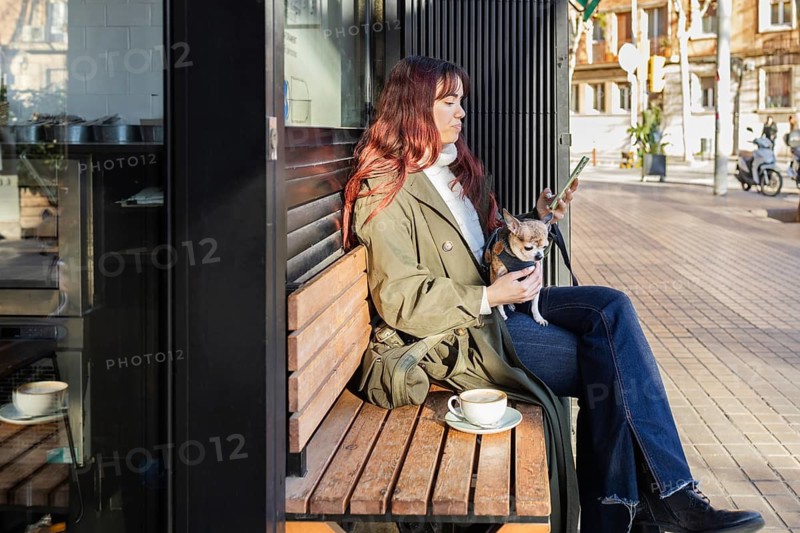A woman sits on a bench outside a cafe, looking at her phone, with a small chihuahua on her lap. She wears a green coat and jeans, while a coffee cup rests nearby