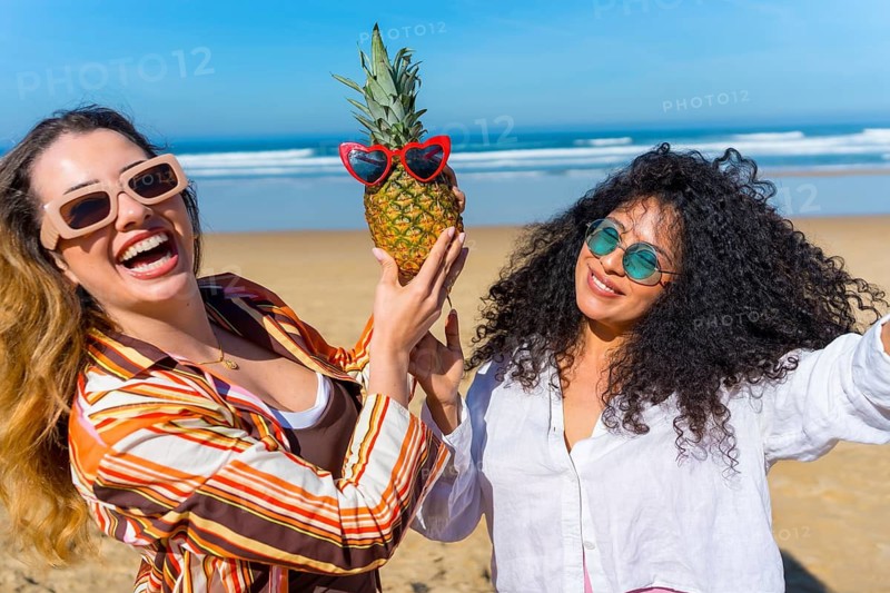 Two young women are smiling and having fun together on a sunny beach, holding a tropical pineapple wearing heart shaped sunglasses, embodying joy and friendship during their summer holiday
