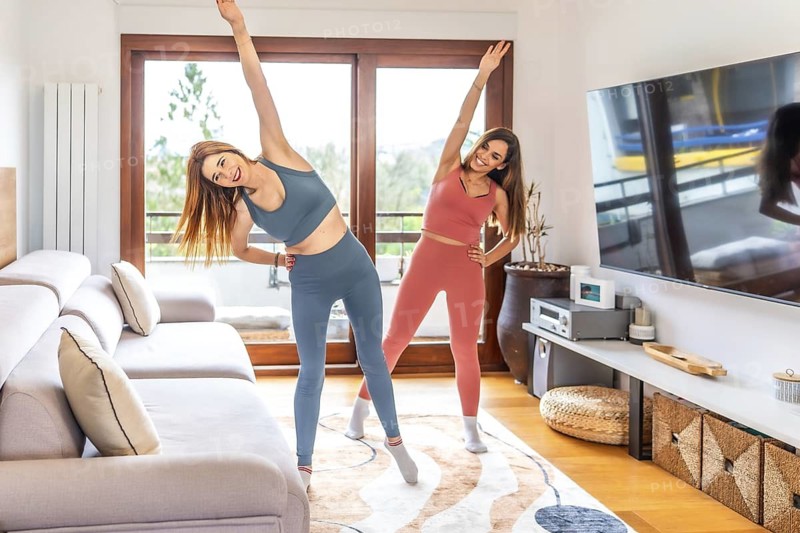 Two smiling women practicing yoga together in their living room, following an online class on their tv