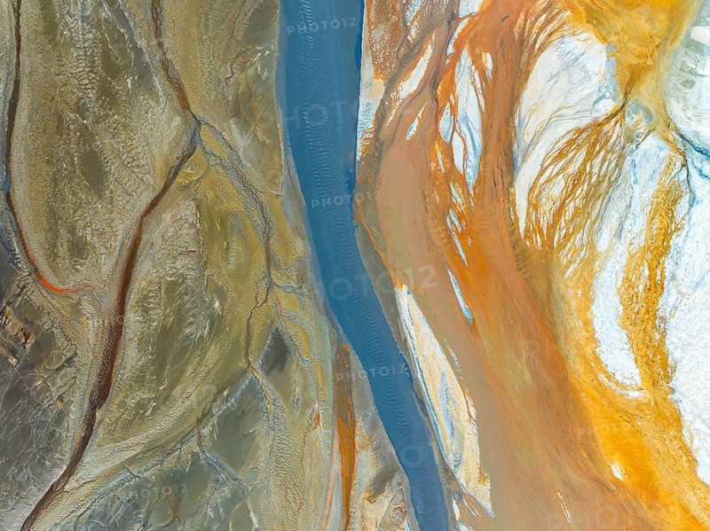 Top view of a vividly colored river meandering through diverse textured terrains, captured by a drone. The contrasting colors and patterns create an abstract nature scene, highlighting the beauty of natural earth formations from above