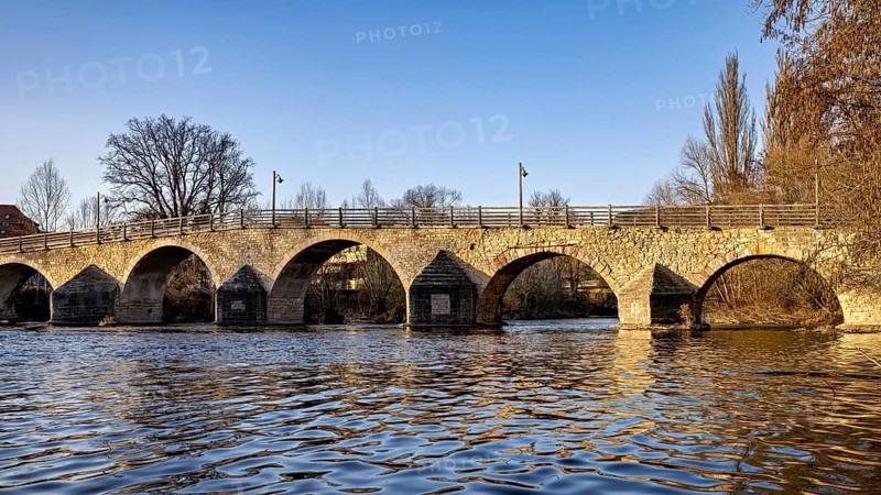 Sunny day view of a stone bridge over calm water, surrounded by trees, The Burgau Bridge in the Saale Valley near Jena in Thuringia