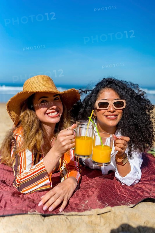 Two smiling friends lying on a blanket at the beach, holding glasses with orange juice, celebrating their summer vacation together under a bright blue sky