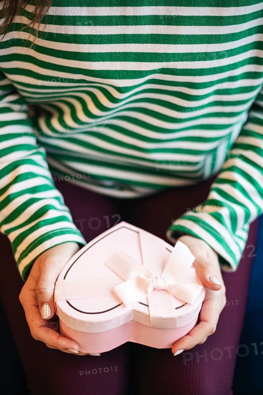 Cropped unrecognizable woman at a cafe holding a heart-shaped gift box decorated with a white ribbon. The image focuses on the gift in her hands with a backdrop of her green and white striped shirt