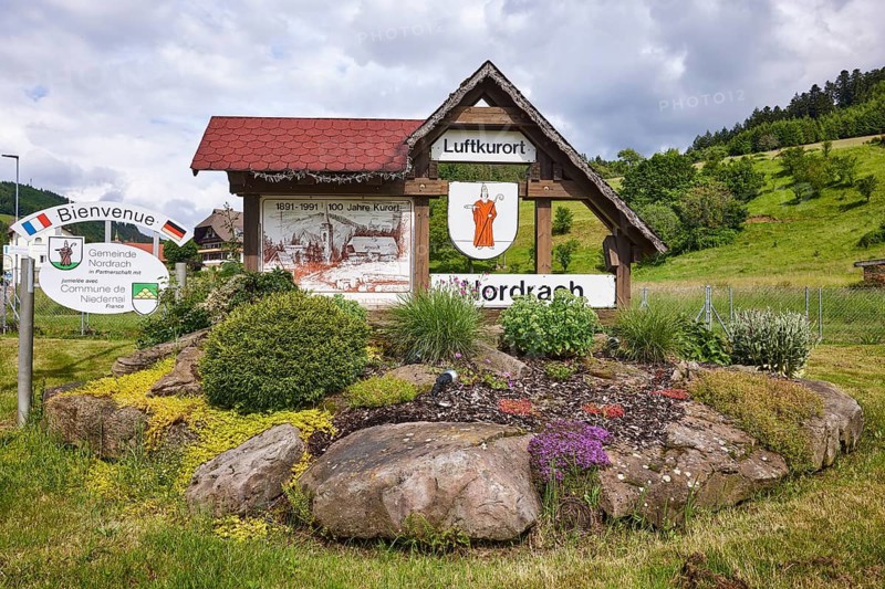 Tourist information sign climatic health resort, flower bed with natural stones, hilly landscape, forest, meadow, village entrance, overcast sky with nimbostratus clouds, street Im Dorf, Nordrach, Black Forest, Ortenaukreis, Baden-Württemberg, Germany