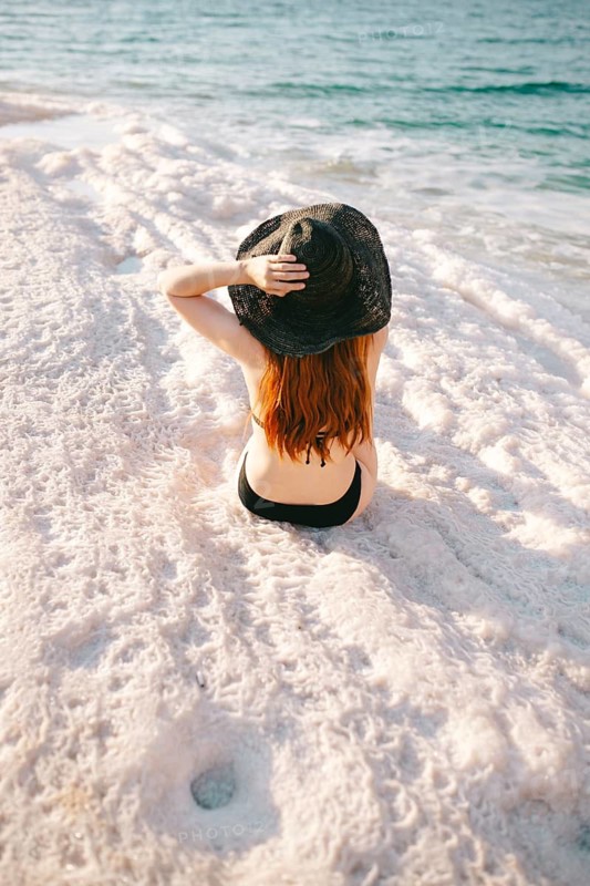 Back view of unrecognizable redhead woman wearing a black hat sitting on the salt formations of the Dead Sea in Jordan, enjoying the view