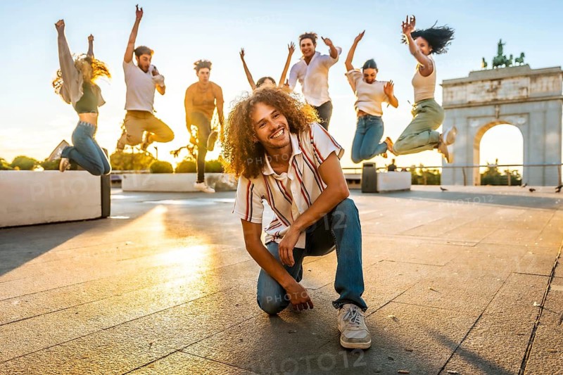 Free style dancer kneeling smiling at camera supporting by multi-ethnic friends jumping on the background in the city during sunset