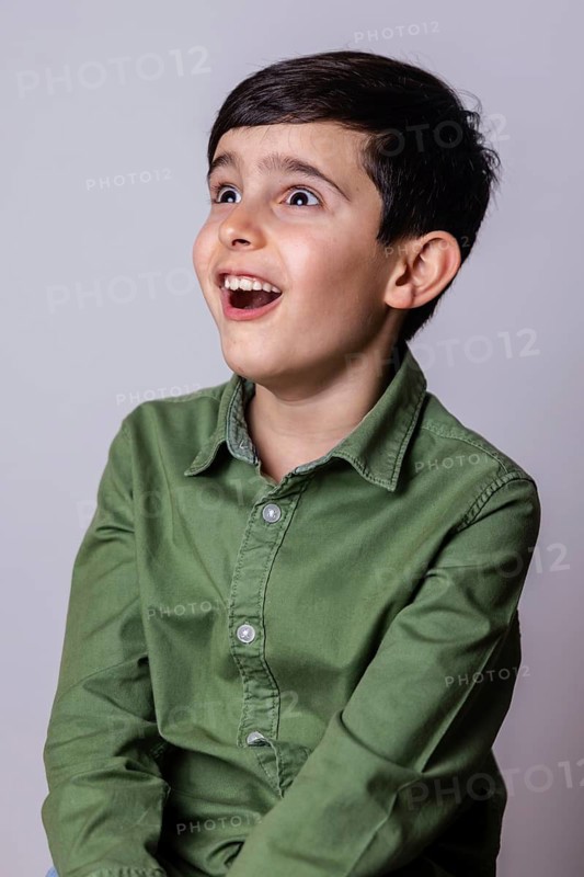 A joyful young boy in a green shirt expresses delight with wide eyes and an open mouth, capturing a moment of pure excitement and happiness against a plain background